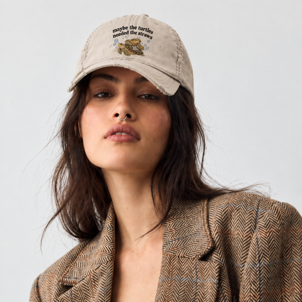 A woman wearing a beige baseball cap with turtles and text that says maybe the turtles needed the straws and a brown herringbone blazer over a white lace top, photographed against a light gray studio background.