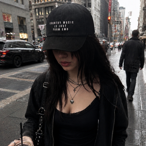 A woman walking on a city street wearing a black baseball cap with white embroidered text that reads 'COUNTRY MUSIC IS JUST FARM EMO'. She is dressed in black with layered necklaces and holds an iced coffee.