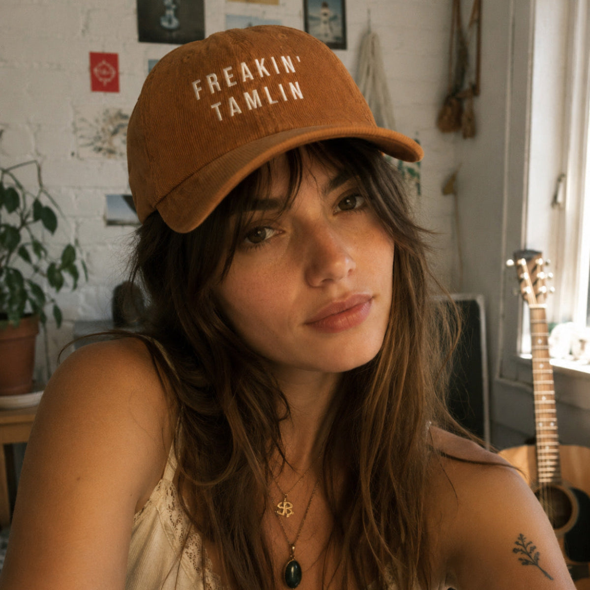 A woman wearing a brown cotton baseball cap with the text 'FREAKIN' TAMLIN' in white, photographed in a warm indoor setting with a guitar and plant in the background.