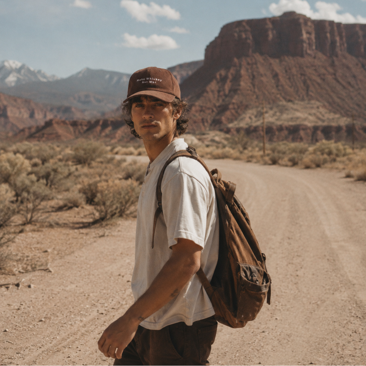 A man wearing a brown baseball cap, white t-shirt, and brown backpack stands on a dirt road in a desert landscape with red rock formations and mountains in the background.